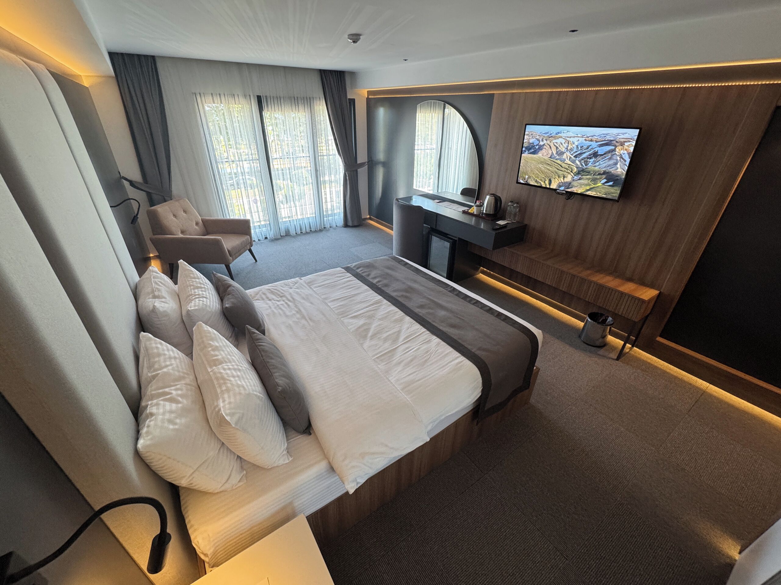 Ein modernes Hotelzimmer mit einem großen Bett, einem Schreibtisch, einem Flachbildfernseher, einem Sessel und Blick ins Freie durch große Fenster.
