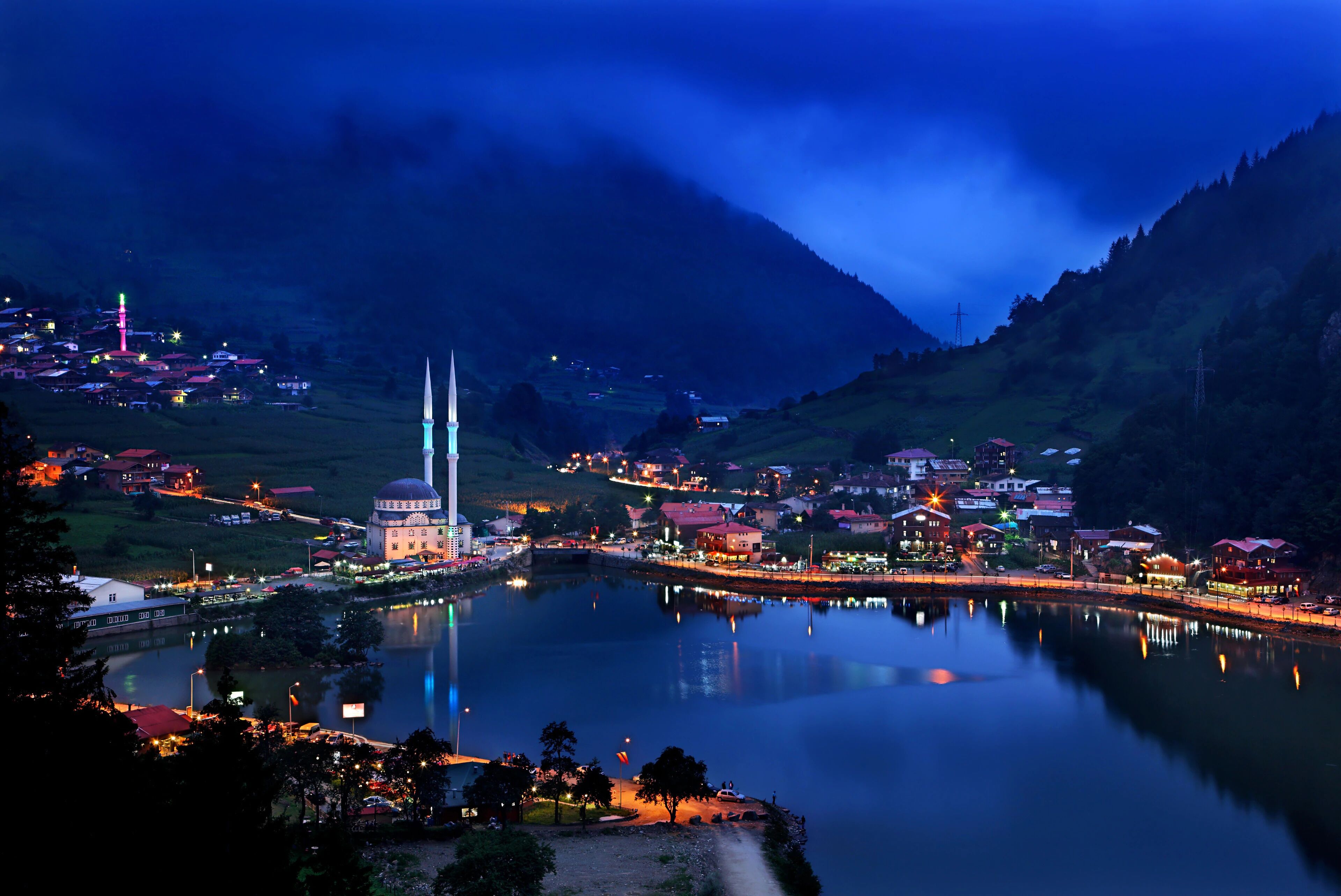 A serene lakeside town at dusk with a prominent mosque, a bridge, and surrounding buildings.