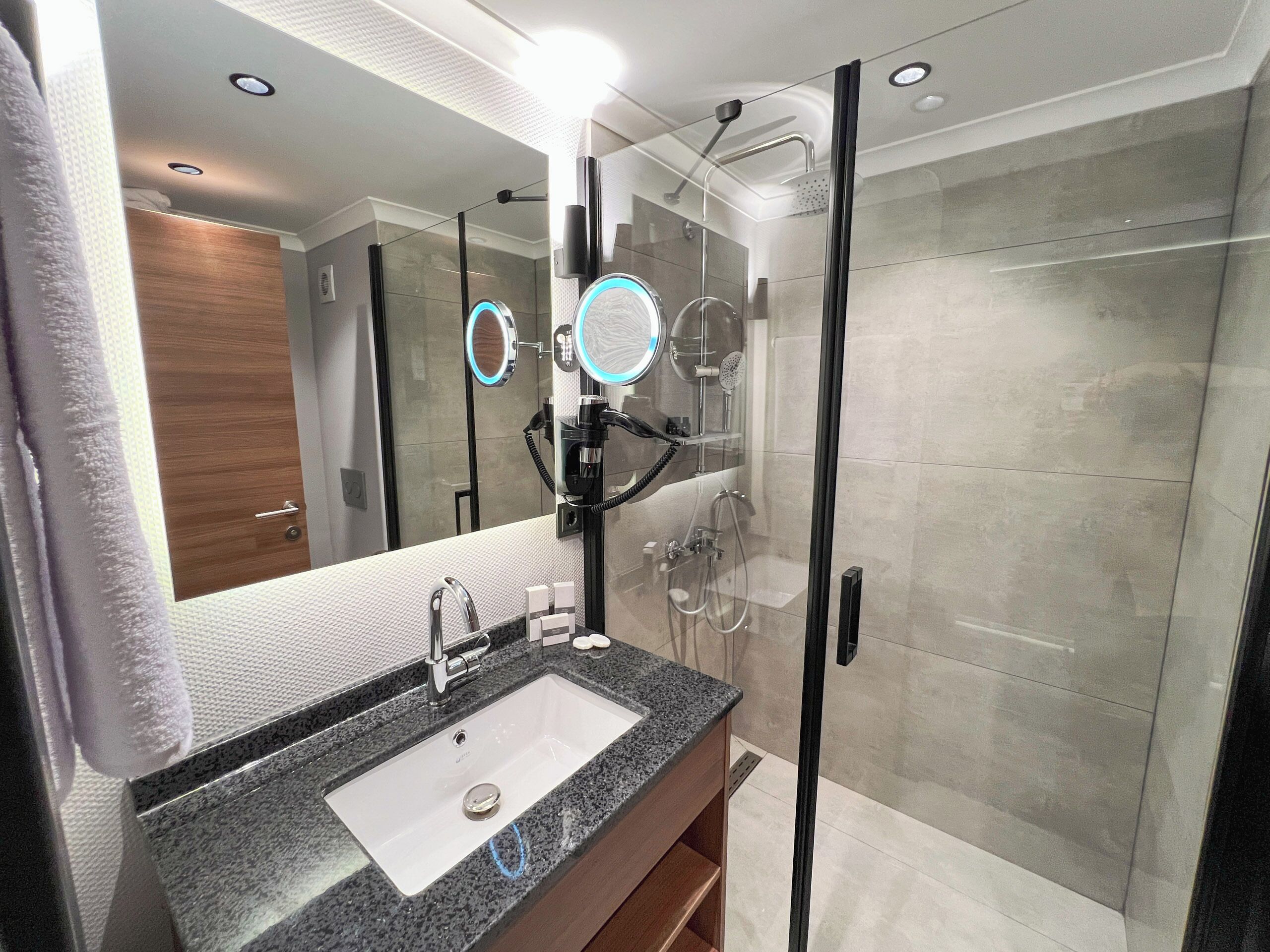 Un baño moderno con lavamanos de granito, cabina de ducha con paredes de vidrio y un espejo grande con iluminación integrada.