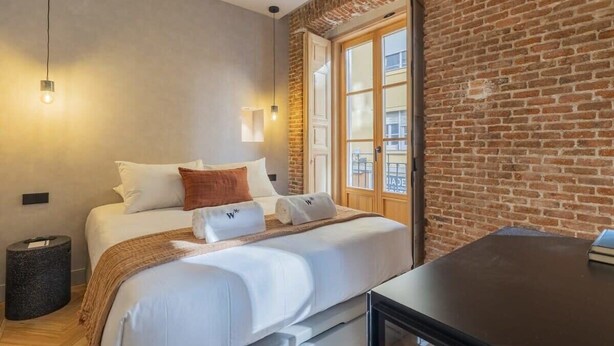 A modern hotel room with a brick wall, a large bed with white linens, a small bedside table, and a window with a view of the city. - w *| Luxury 1BR at Palacio Real (Madrid)