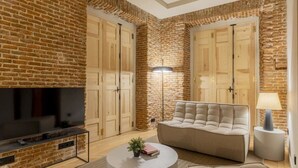 A living room with a brick wall, a flat-screen TV, a beige sofa, a round coffee table, and a lamp. - w *| Luxury 1BR at Palacio Real (Madrid)