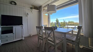 Une pièce avec un téléviseur posé sur un meuble blanc, une table à manger avec des chaises et une grande fenêtre donnant sur un paysage verdoyant.