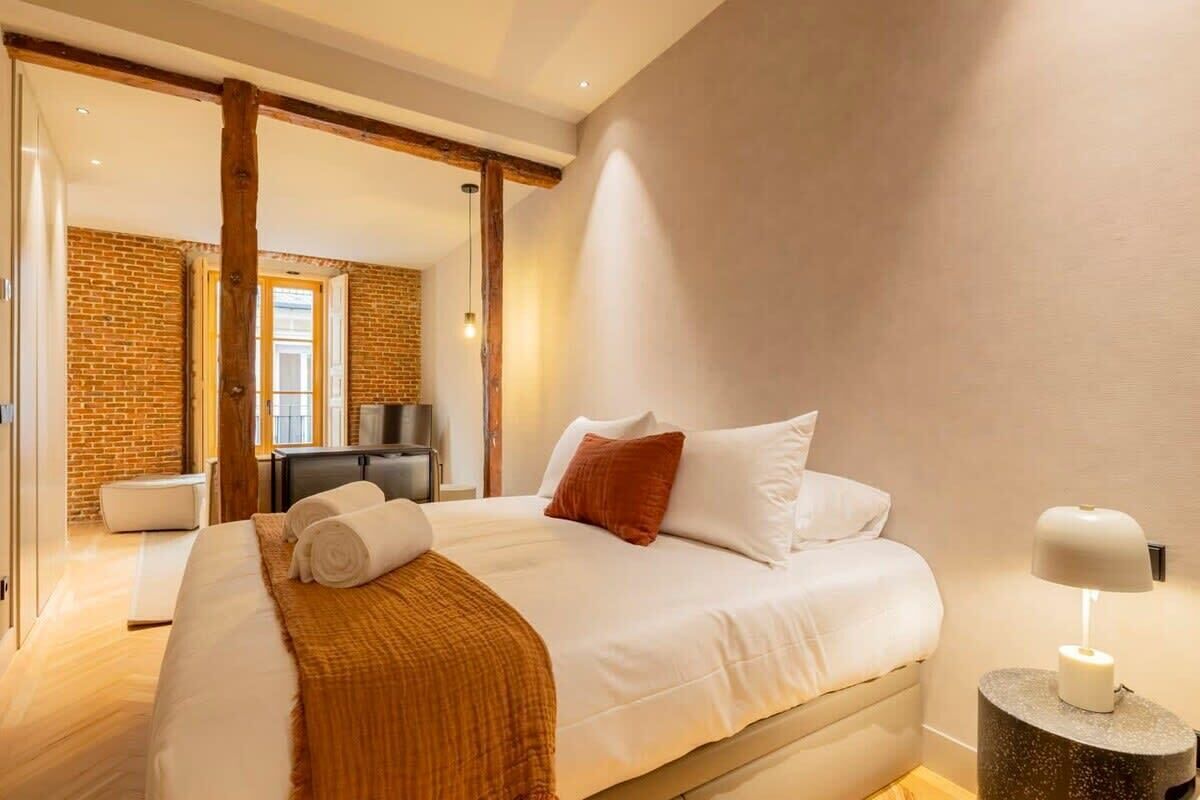 Un dormitorio moderno con una cama grande, techo de vigas de madera, pared de ladrillo y una mesita de noche con lámpara.