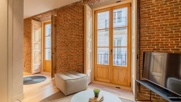 A modern living room with exposed brick walls, a large window, a sofa, a coffee table, and a TV. - w *| Amazing 1BR at Palacio Real (Madrid)