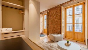 A modern hotel room with a large window, a brick accent wall, a sofa, a small table, and a wardrobe. - w *| Amazing 1BR at Palacio Real (Madrid)