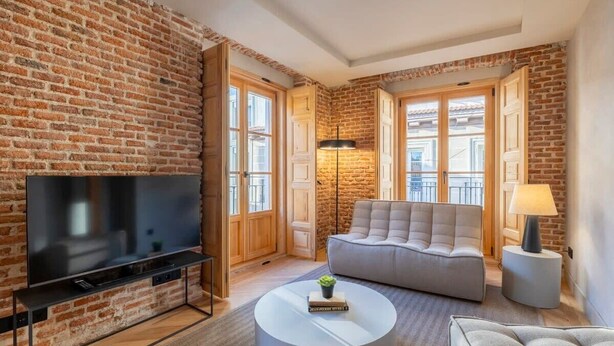 A living room with a brick wall, a flat-screen TV, a grey sofa, a round coffee table, and a lamp. - w * | Modern 1BR at Palacio Real (Madrid)