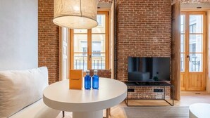 A modern living room with a brick wall, a round white table with a lamp, a sofa, and a flat-screen TV on a brick fireplace. - w * | Modern 1BR at Palacio Real (Madrid)