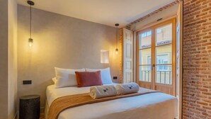 A hotel room with a large bed, a small round stool, a window with a view of buildings, and a brick wall. - w * | Modern 1BR at Palacio Real (Madrid)