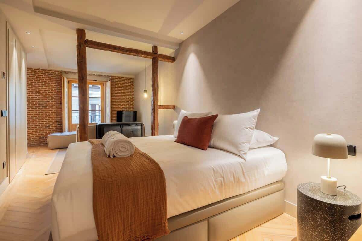 Une chambre d’hôtel moderne avec un grand lit, une poutre en bois au plafond, un mur en briques et un petit bureau avec une lampe.