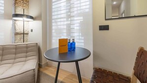 A modern living room with a sofa, a round table with a book and bottles, a lamp, and a brick wall. - Cozy 1BR at Palacio Real (Madrid)