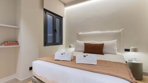 A neatly made bed with white linens and two towels with a logo, a bedside table with a lamp, and a window with blinds. - Cozy 1BR at Palacio Real (Madrid)