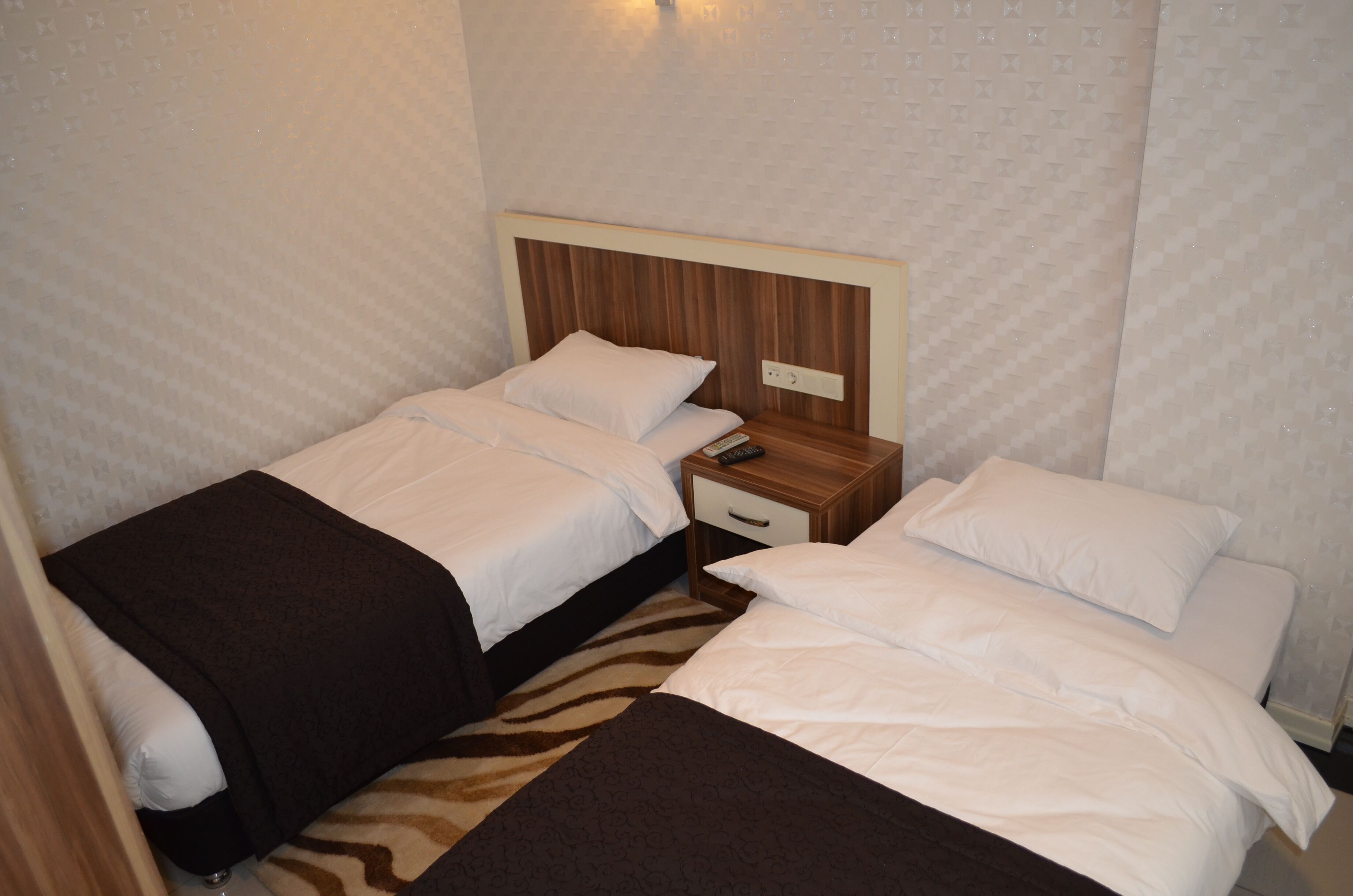 A hotel room with two single beds, a wooden nightstand with a drawer, and a wall-mounted light fixture.