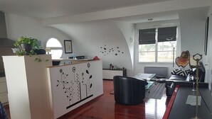 Ein modernes Wohnzimmer mit Holzboden, einer schwarzen Ledercouch, einem Schreibtisch mit Computer und einer Zimmerpflanze auf einem weißen Schrank.