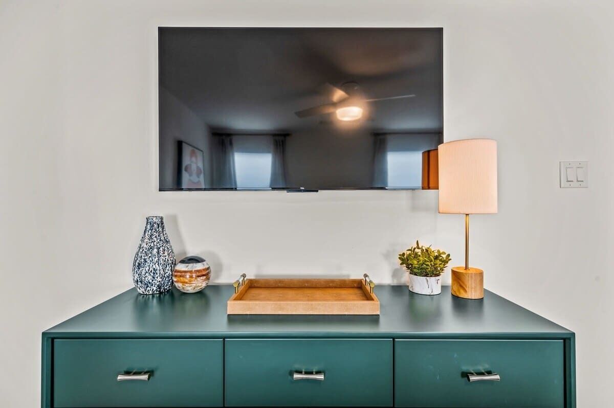 A teal-colored dresser with a flat-screen TV mounted above it, a wooden tray, a vase, a lamp, and a small potted plant.