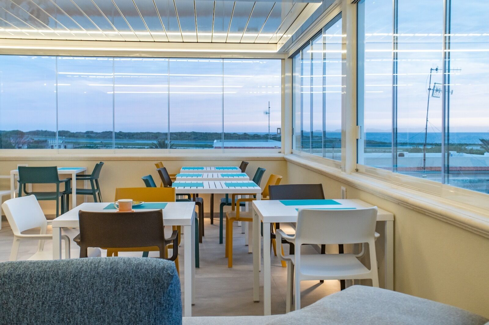 Ett modernt inomhusutrymme med bord och stolar arrangerade för måltider eller möten, stora fönster med utsikt över flygplatsen och landningsbanan, och ett tak med linjära ljusarmaturer.