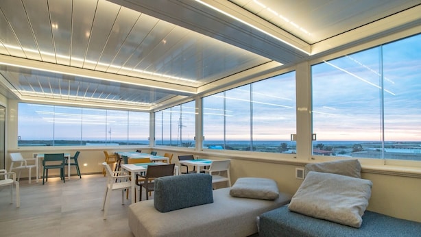 A spacious room with a large window offering a view of the sea and cityscape. There are several seating areas, including a sofa and chairs, arranged around a central table.