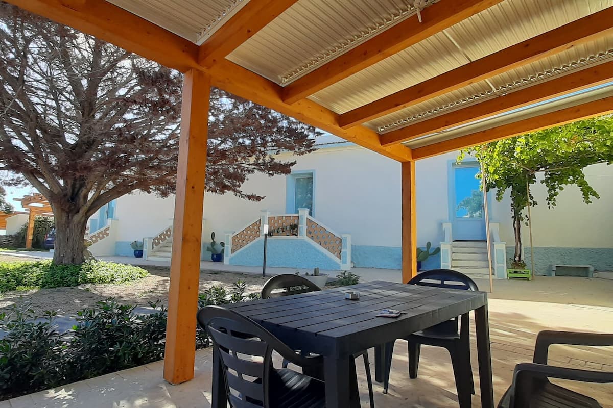 An outdoor patio area with a wooden pergola, a table with chairs, and a staircase leading to a building with blue and white walls.
