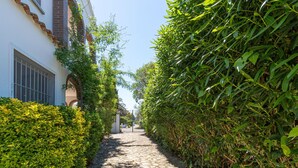 A narrow cobblestone path flanked by dense green hedges and a white building with a brick facade and barred windows. - "Holiday Home for 6 with Pool, BBQ, Wifi, Near Beach - Sant Pere Pescador, Costa Brava" (Sant Pere Pescador)