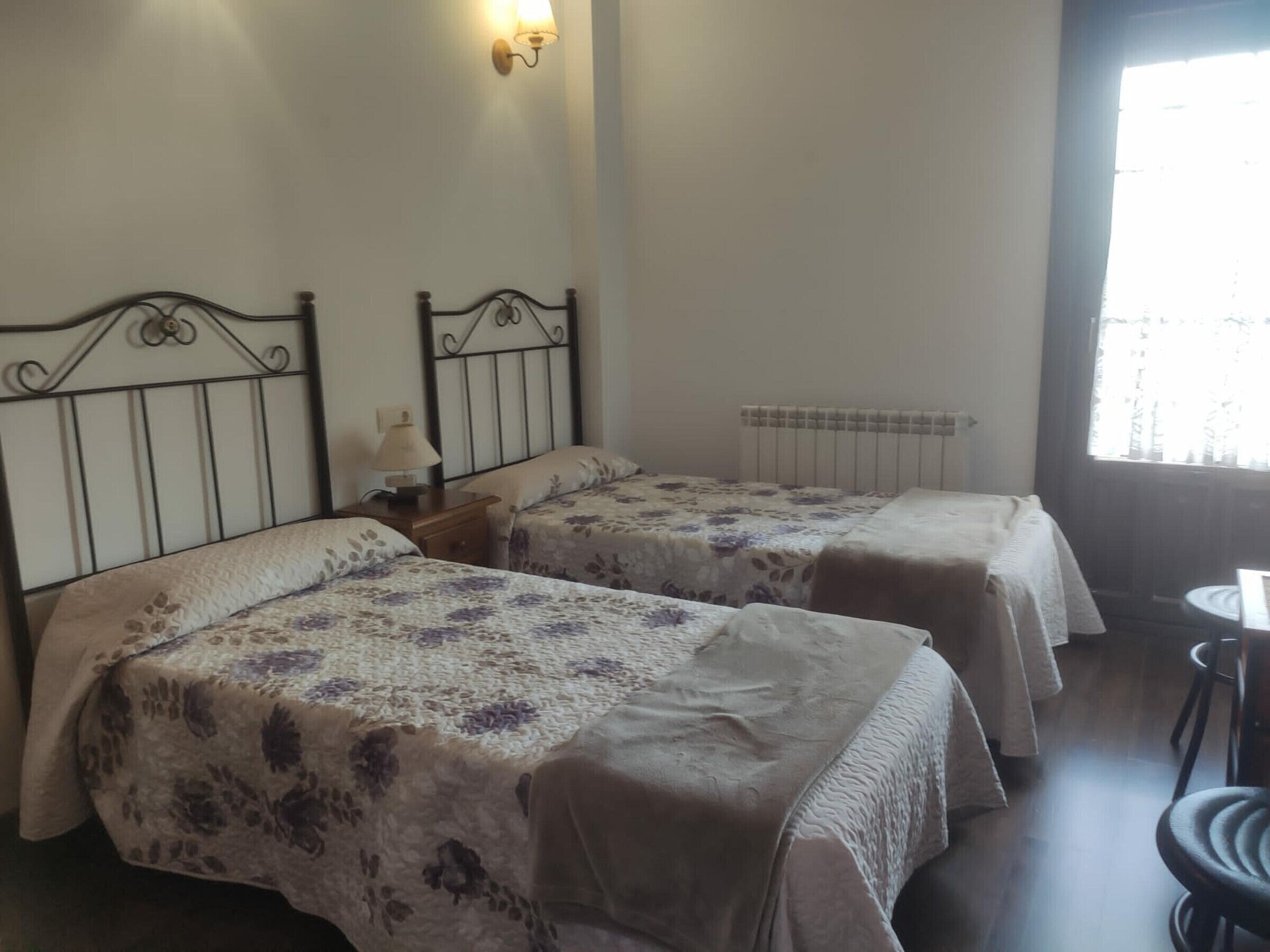 A room with two beds, a nightstand with a lamp, a radiator, and a window with a view of trees.