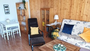 A living room with a wooden floor, a black chair with a yellow cushion, a white sofa with blue and yellow cushions, a wooden coffee table with a decorative bowl, a white table with chairs, and a wooden cabinet with shelves. - Spacious 2-bedroom apartment with parking in Villanúa (Villanúa)