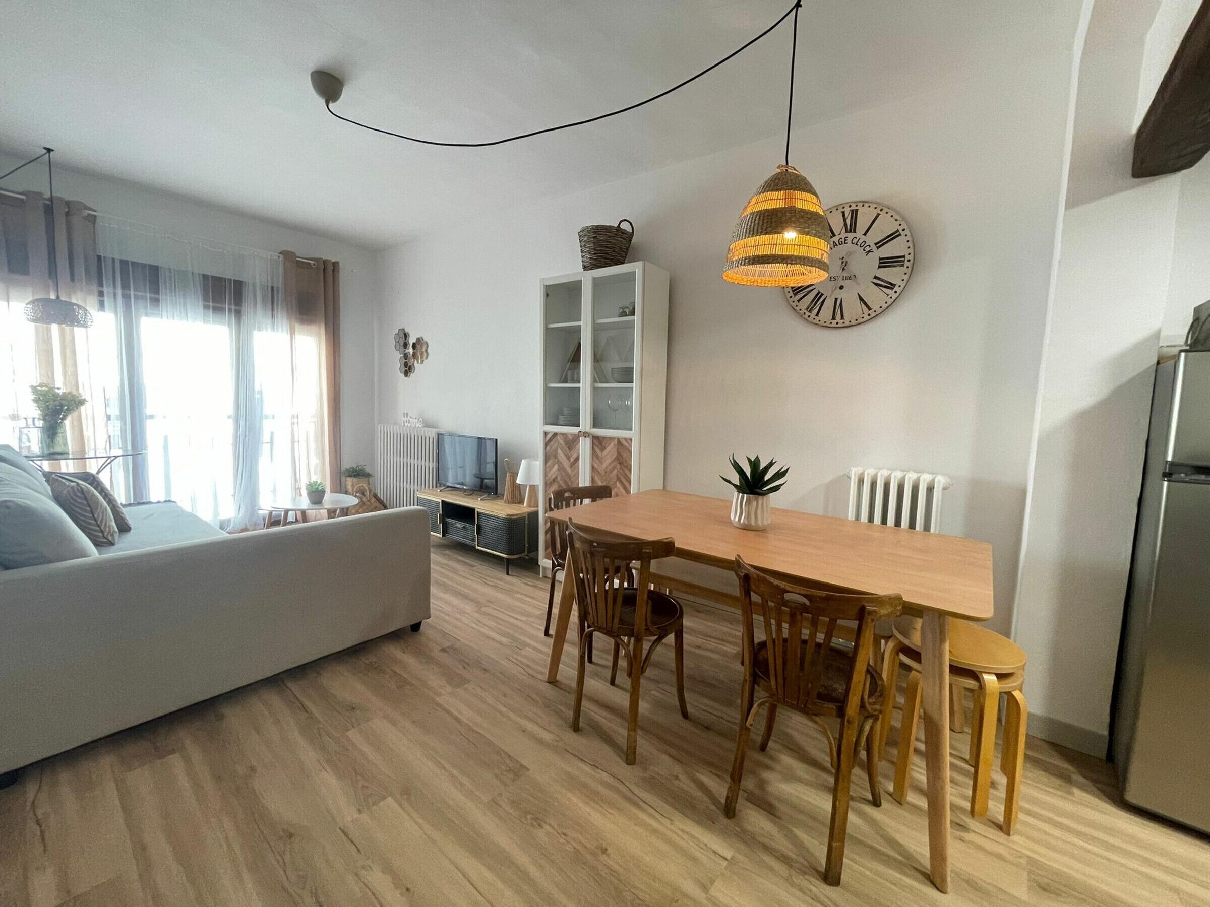 A modern living room with a sofa, dining table, and chairs. There is a television, a clock, and a radiator.