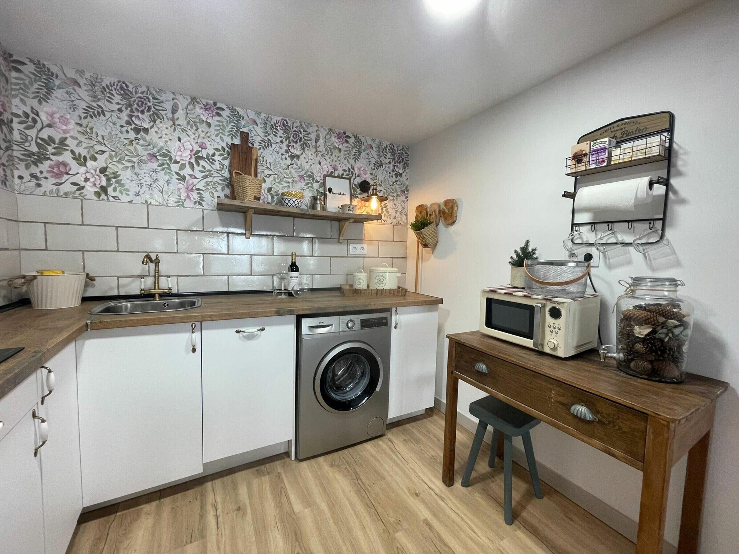 A compact kitchen with a washing machine, a microwave, a small wooden table, and a shelf with kitchen items.