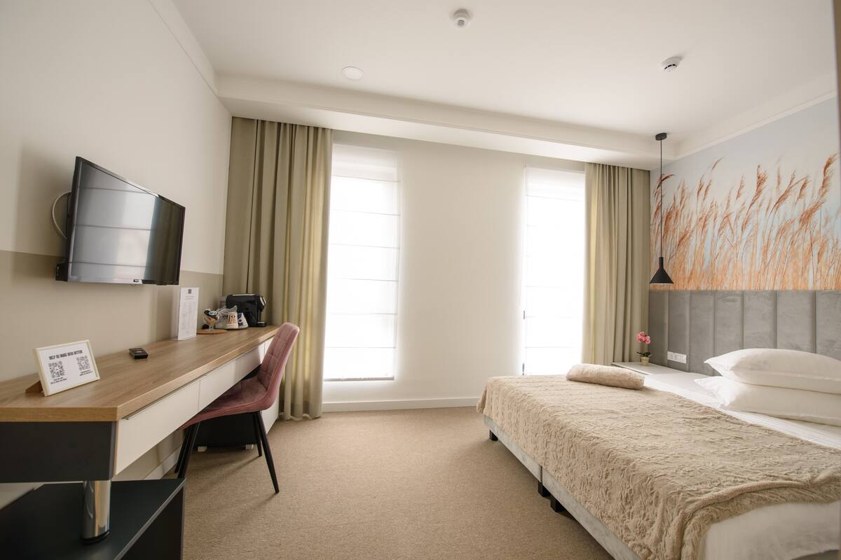 A hotel room with a bed, a desk with a chair, a television, and a large window with curtains.