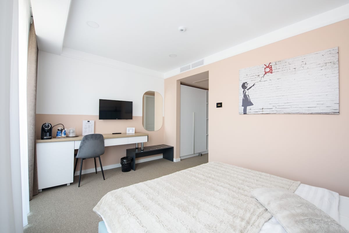 A hotel room with a bed, a desk with a chair, a television, a mirror, and a painting on the wall.
