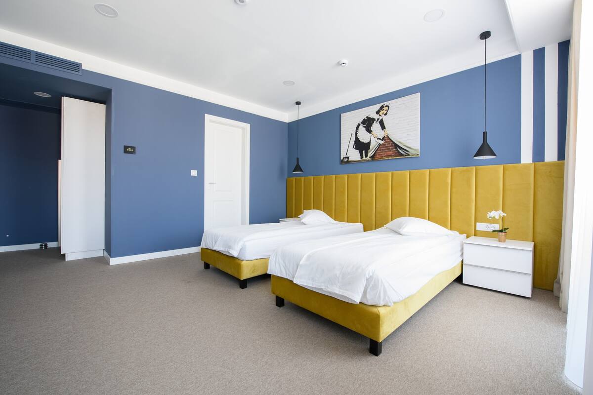 A hotel room with two beds, a yellow headboard, a white nightstand, and a painting on the wall.