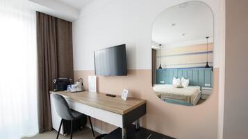 A hotel room with a wooden desk, a black chair, a flat-screen TV, a bed with a blue headboard, and a large mirror.
