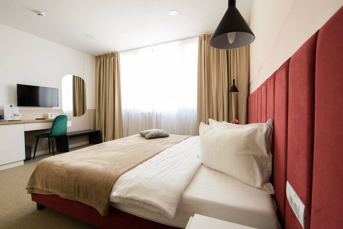 A hotel room with a bed, a desk with a chair, a television, and a window with curtains.