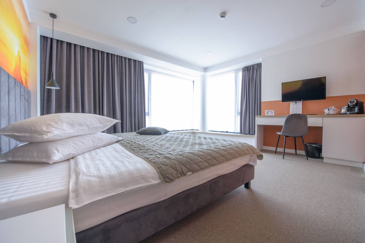 A modern hotel room with a large bed, a desk with a chair, a television, and a window with curtains.