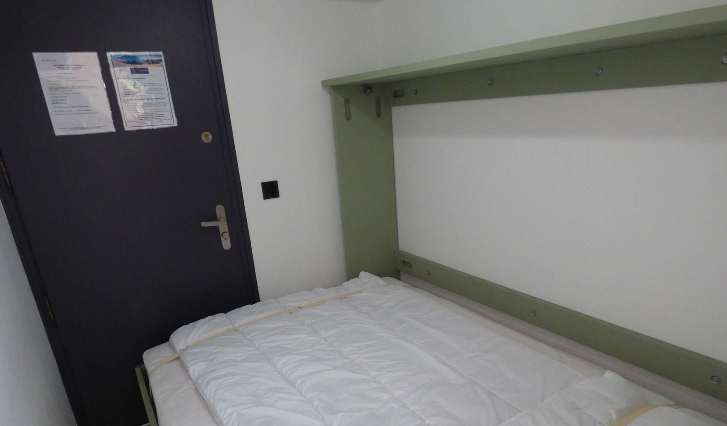 A small, single-bed room with a plain white mattress and a green headboard. There is a door with a lock and two notices attached to it.