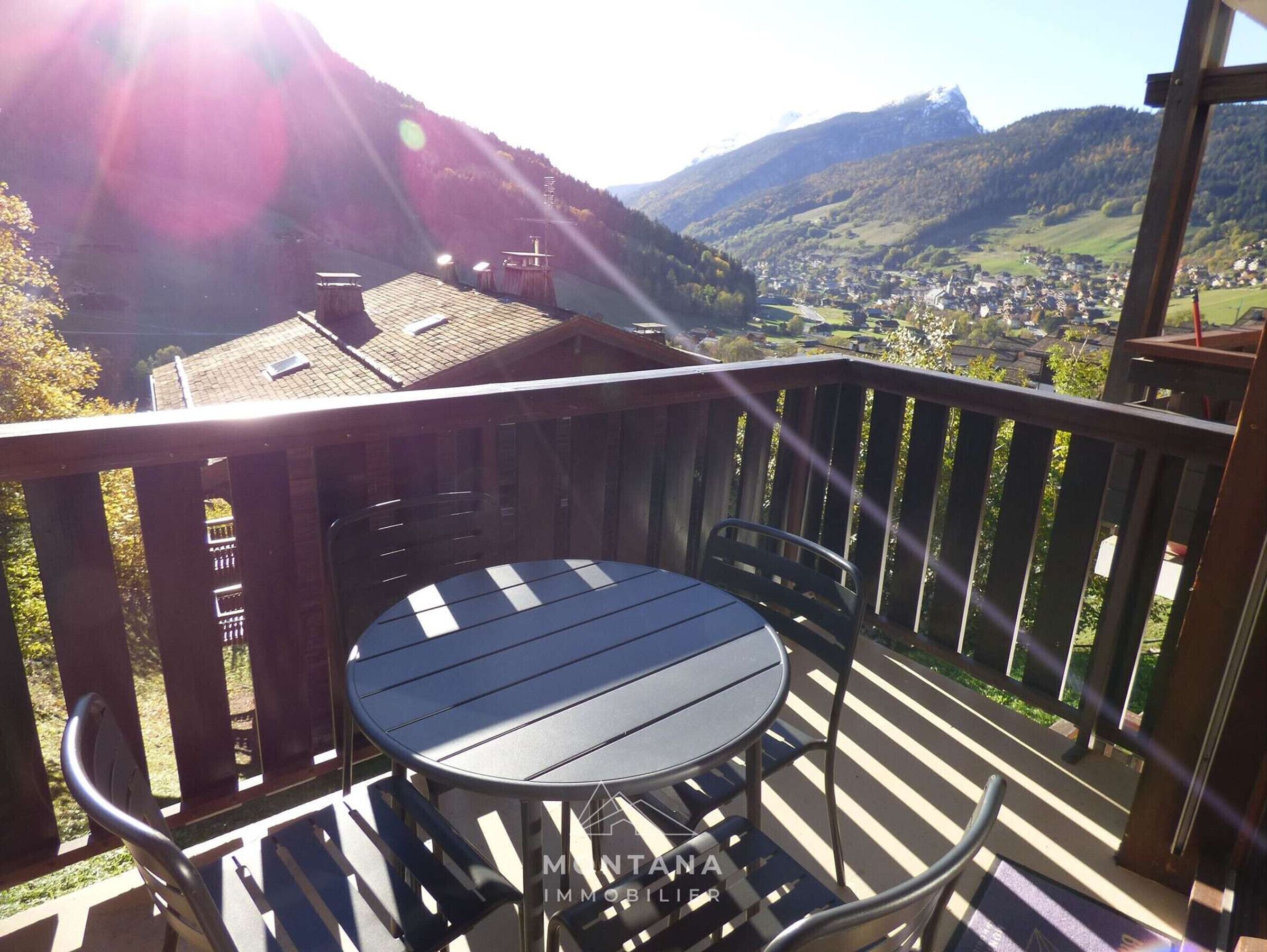 Una terraza de madera con una mesa redonda y sillas, con vistas a un valle con casas y montañas.