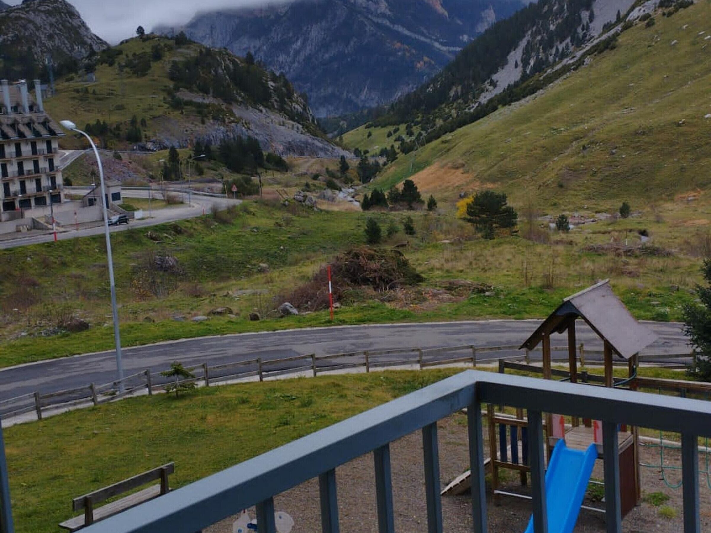 A playground with a slide and a small shelter, surrounded by a road and a mountainous landscape.