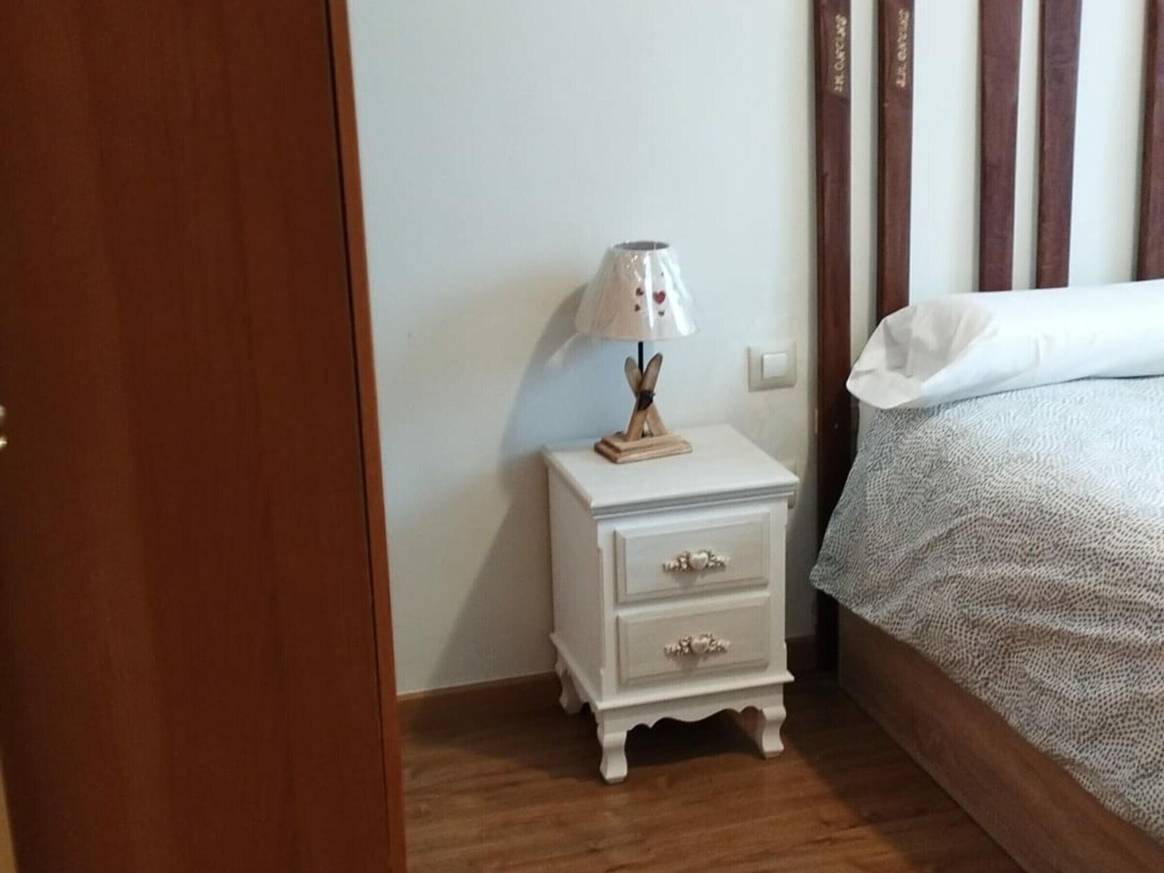 Um quarto com piso de madeira, mesa de cabeceira branca com gavetas, luminária e cama com travesseiro branco e manta texturizada.