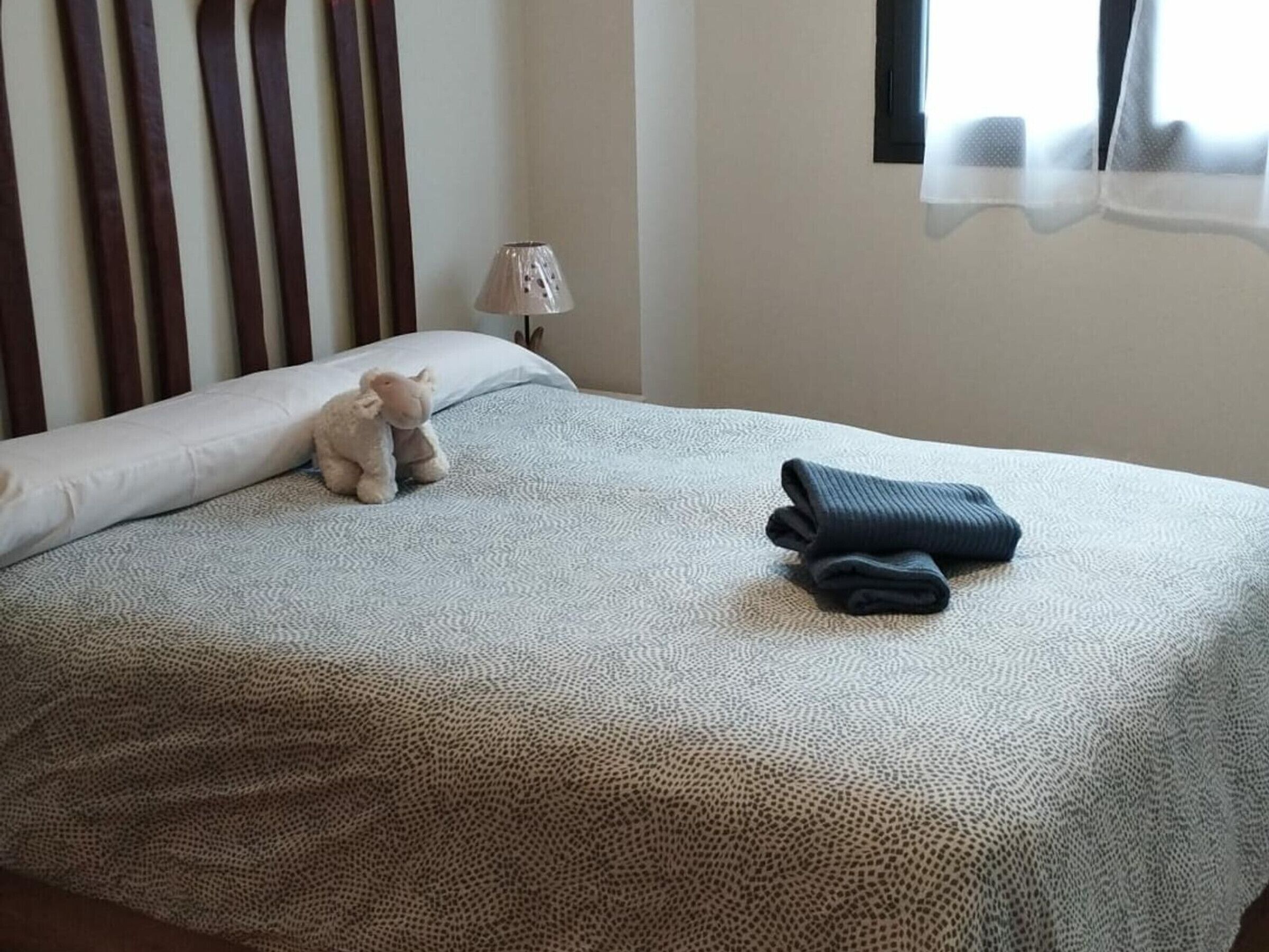 Una cama bien hecha con una colcha estampada, una toalla doblada y un osito de peluche sobre las almohadas.