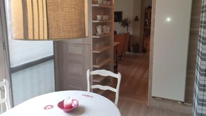Un coin repas avec une table, des chaises et une lampe. On aperçoit également une cuisine avec des placards et un réfrigérateur au fond.