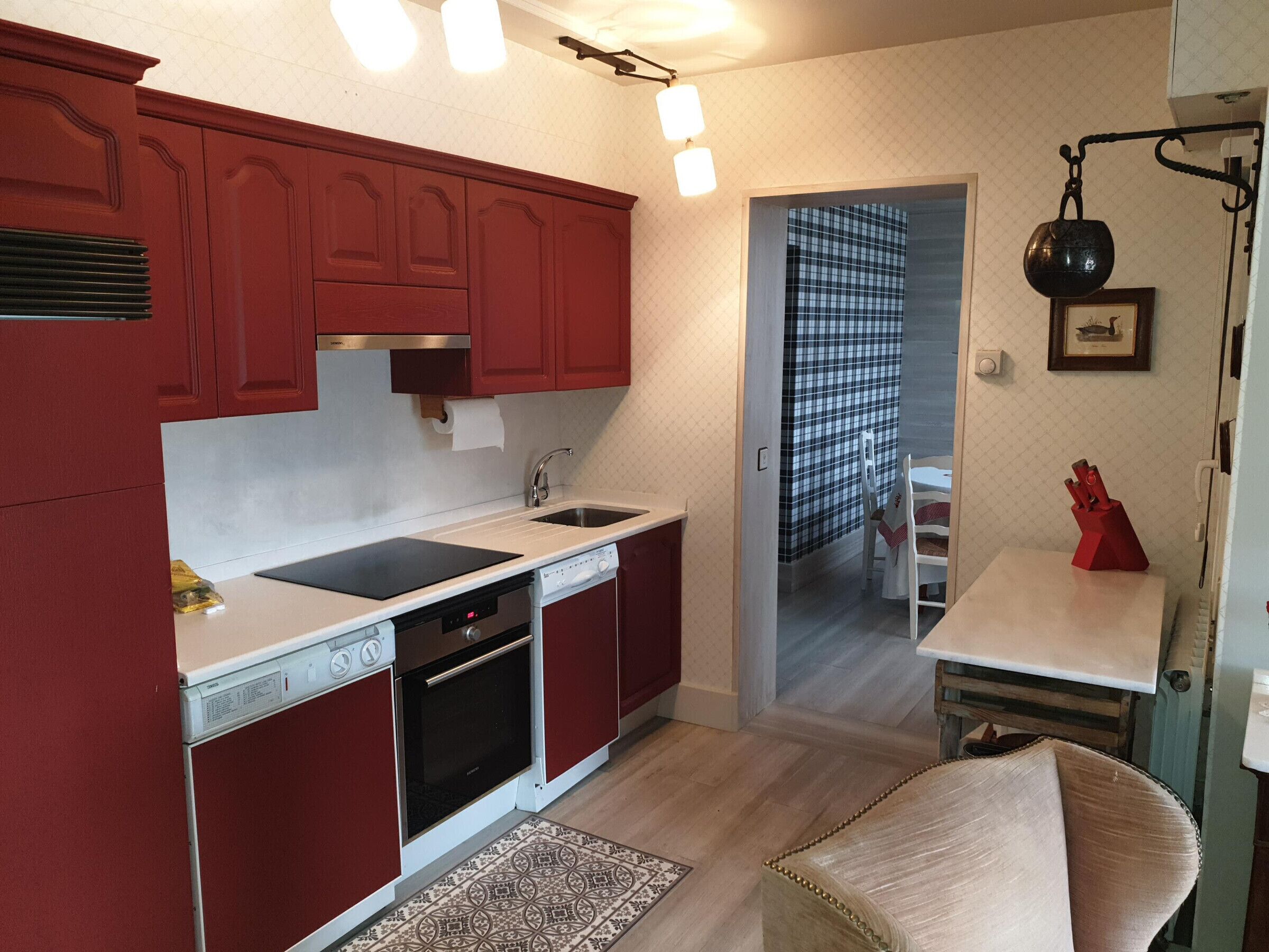 Een kleine keuken met rode kastjes, een wit aanrecht, een ingebouwde oven en een spoelbak. Er is een zithoek met een tafel en stoelen, en een vloerkleed met een patroon.