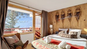 A hotel room with a bed, a chair, a window with a view of a snowy mountain, and wooden wall decor. - Cozy apartment at pistes, near shops (Méribel)