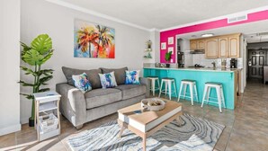 A modern living room with a grey sofa, colorful cushions, a coffee table, and a kitchen area with a turquoise bar and white stools. - Meridian Plaza Unit 1402 - Oceanfront! (Myrtle Beach)