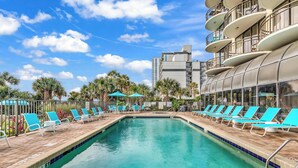 A swimming pool with blue chairs and umbrellas, surrounded by palm trees and a modern building with balconies. - Meridian Plaza Unit 1402 - Oceanfront! (Myrtle Beach)
