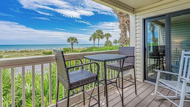 A balcony with wicker chairs and a table overlooking a beach with palm trees and the ocean.