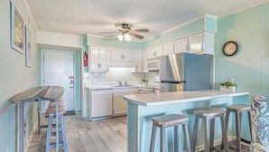 A kitchen with white cabinets, a stainless steel refrigerator, a microwave, and a ceiling fan. There are three bar stools and a small island with stools.