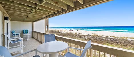 A covered outdoor area with a table and chairs, overlooking a beach with clear blue water and sandy shore.