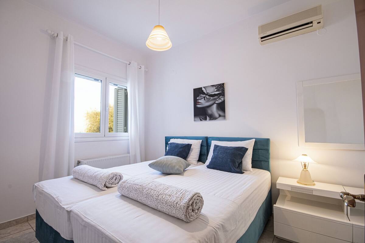 Una cama bien hecha con dos almohadas, una toalla enrollada y una lámpara de noche.
