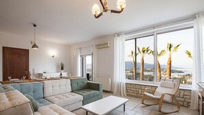 A modern living room with a sectional sofa, a small table, and a rocking chair. There is a large window with a view of palm trees and a distant landscape. - Ionian Star Villas by Corfu Escapes (Corfu)