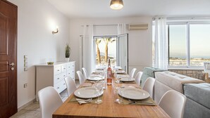 A dining area with a wooden table set for six, white chairs, a sofa with cushions, and a view of palm trees through a window. - Ionian Star Villas by Corfu Escapes (Corfu)