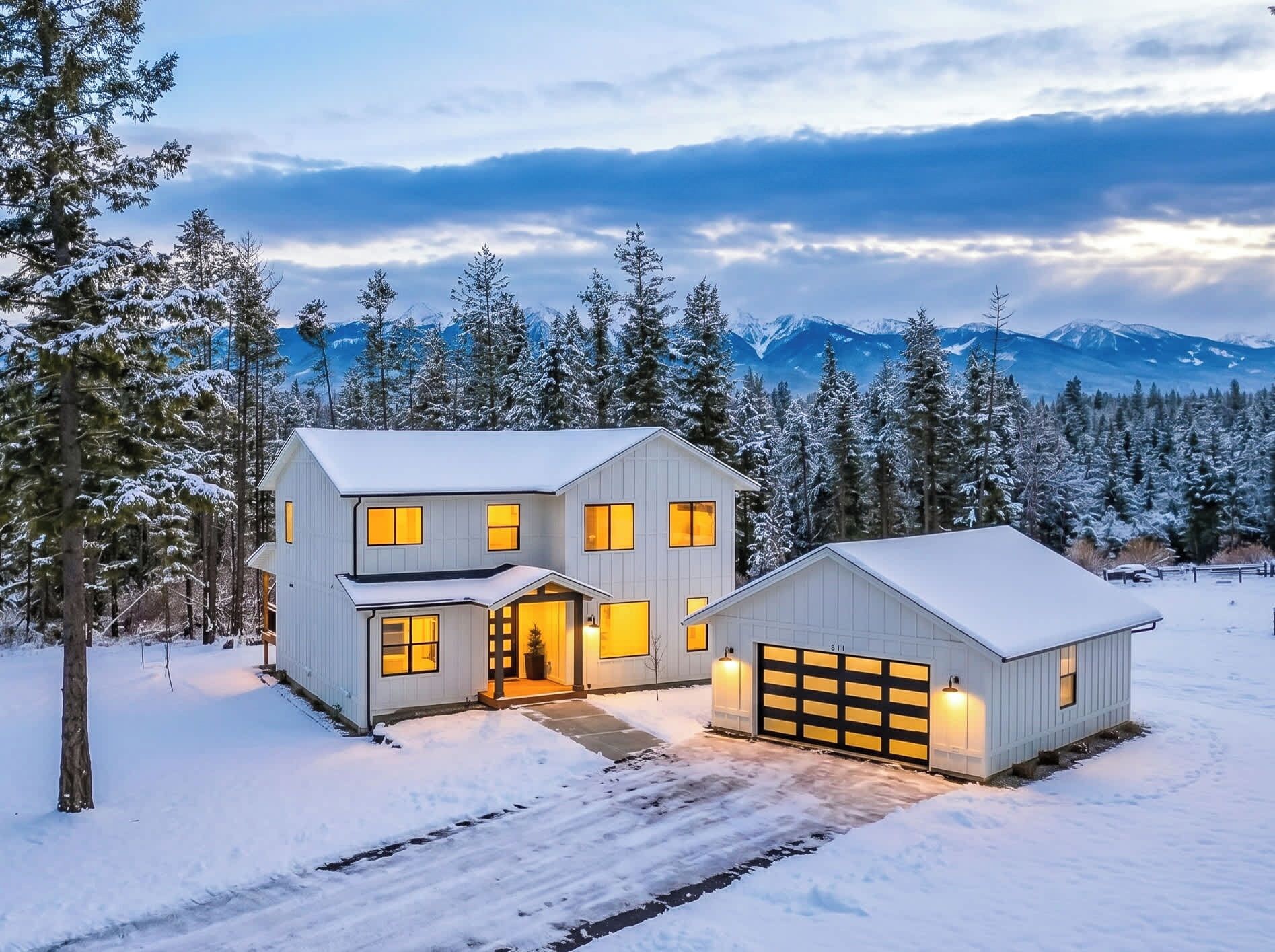 A two-story house with a garage, surrounded by snow-covered trees and mountains in the background.