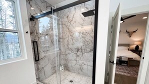 A modern bathroom with a glass shower enclosure, marble walls, and a visible bedroom with a bed and a wall-mounted bull skull decoration.